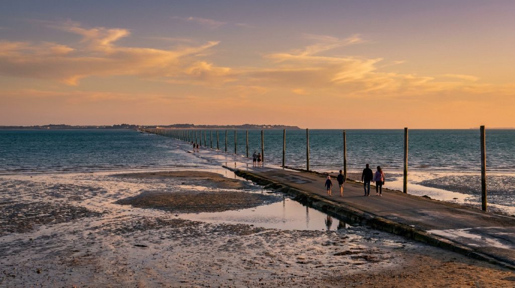 Passage du Gois à Noirmoutier au coucher du soleil. La chaussée pavée, entourée de l'océan et de la grève, est empruntée par des marcheurs.