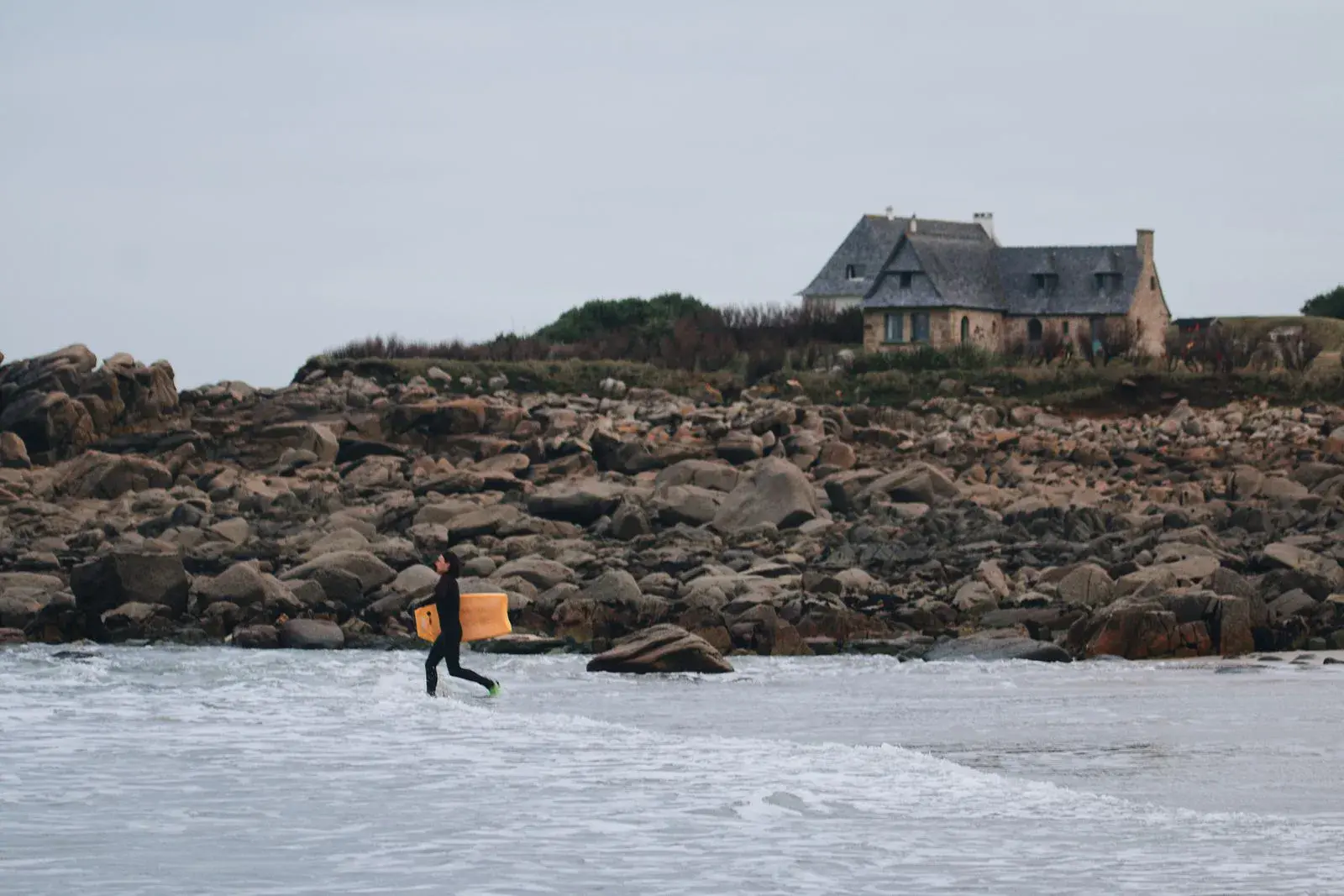 Bodyboarder en combinaison intégrale entrant dans l'eau avec sa planche, devant une côte rocheuse de granit par temps gris