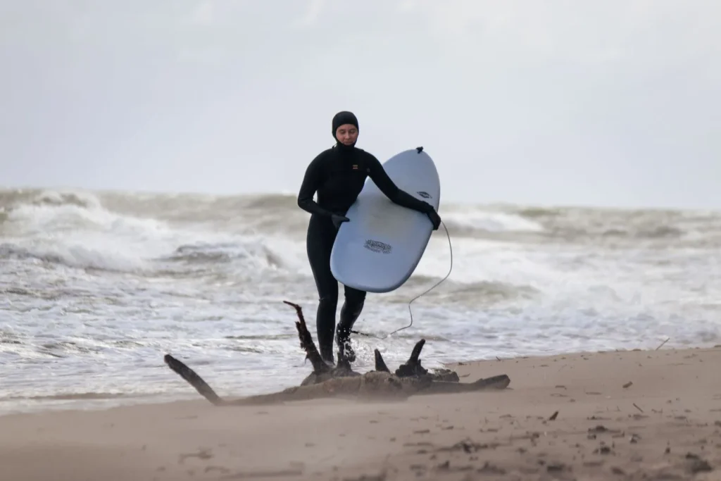 Surfeur en combinaison d'hiver à cagoule et gants sortant de l'eau sur une plage venteuse